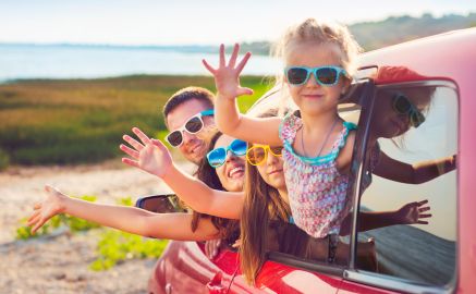 Ein Familie mit bunten Sonnenbrillen auf der Nase winkt aus den Fenstern eines roten Autos.