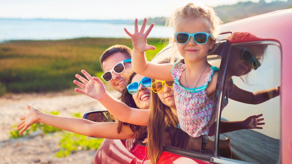 Ein Familie mit Sonnenbrillen winkt aus  dem Auto
